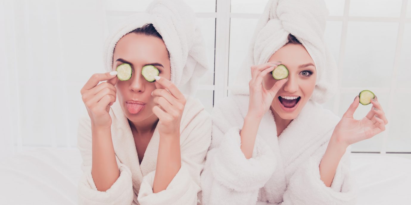 Funny young girls in bathrobes making mask with cucumber and having fun