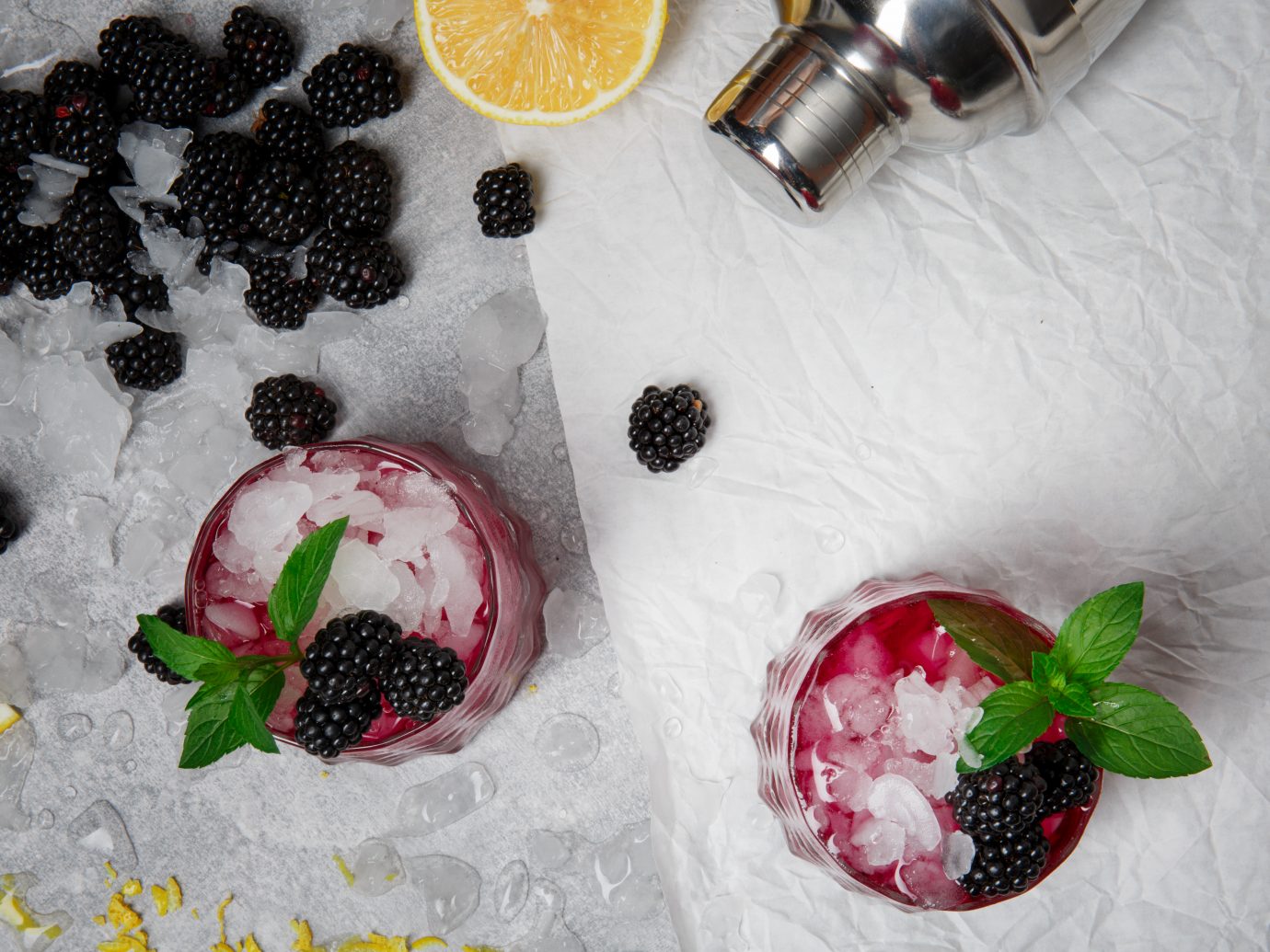 A view from above on a composition of two glasses of icy beverages with peppermint and blackberries on a frozen white background. Ripe, fresh blackberries and a cut yellow lemon for a cold smoothie.