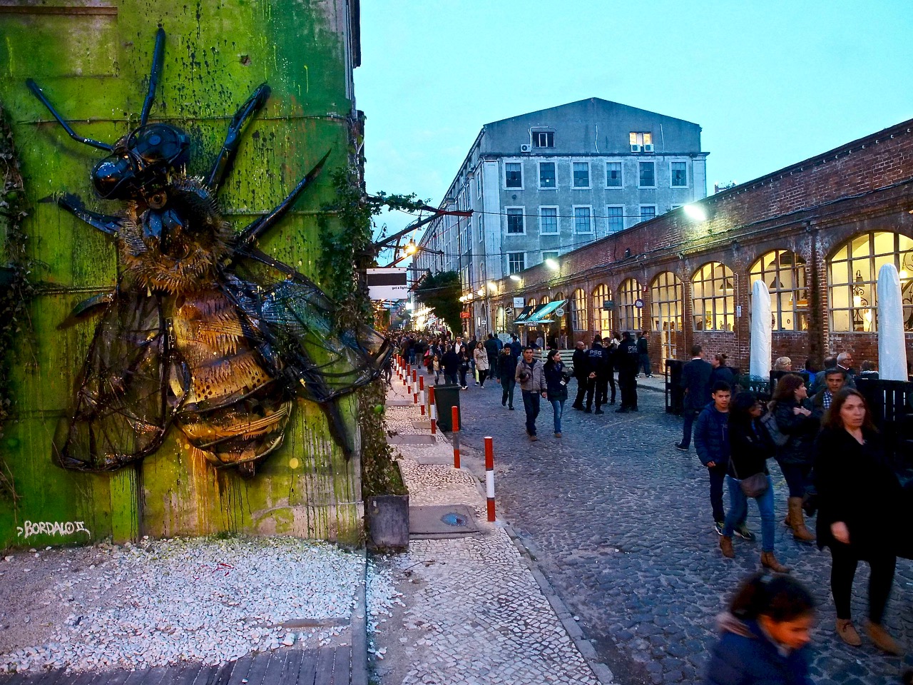 Wall art with large bumble bee and a street with tons of people at dusk at LX Factory