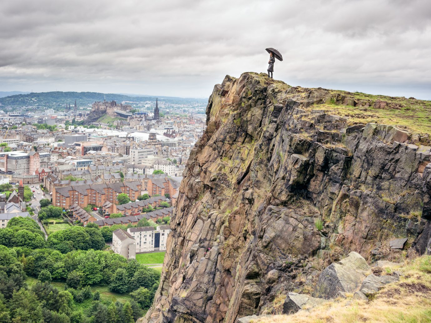 A woman standing above Salisbury Crags, in Holyrood Park, holding an umbrella for shelter as she looks over the city of Edinburgh.