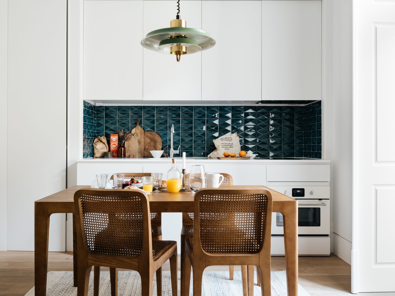 Kitchen interior with basket chairs, a set breakfast on the table, and teal tiling with white cabinets