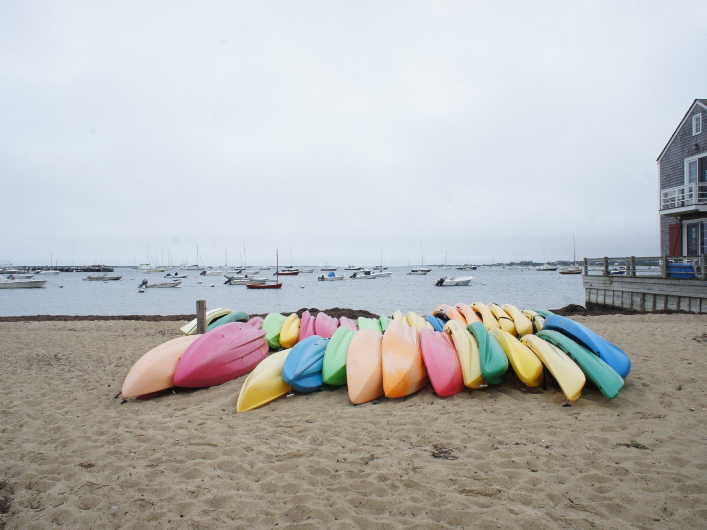 Canoes of assorted colors stacked on one another on a bay beach on a dreary day in Nantucket