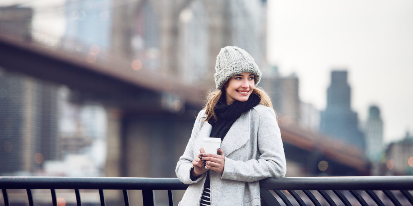 Happy smiling adult tourist woman holding paper coffee cup and enjoying the New York City view and Brooklyn bridge