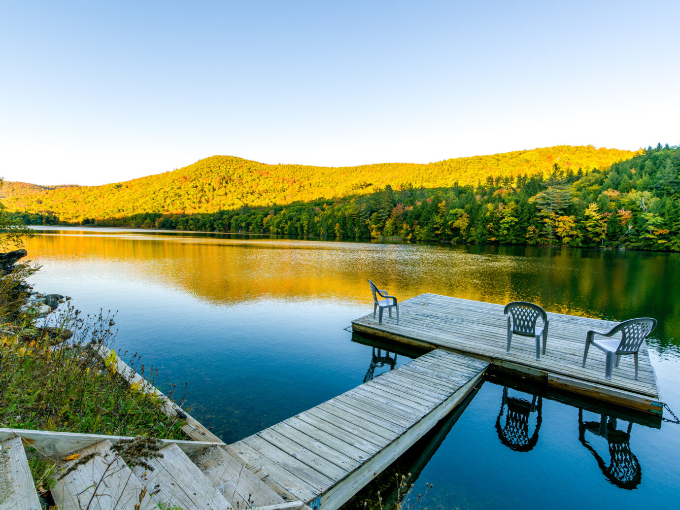 A small deck in the middle of a lake in Vermont. Three chairs are on the deck, inviting people to have a rest